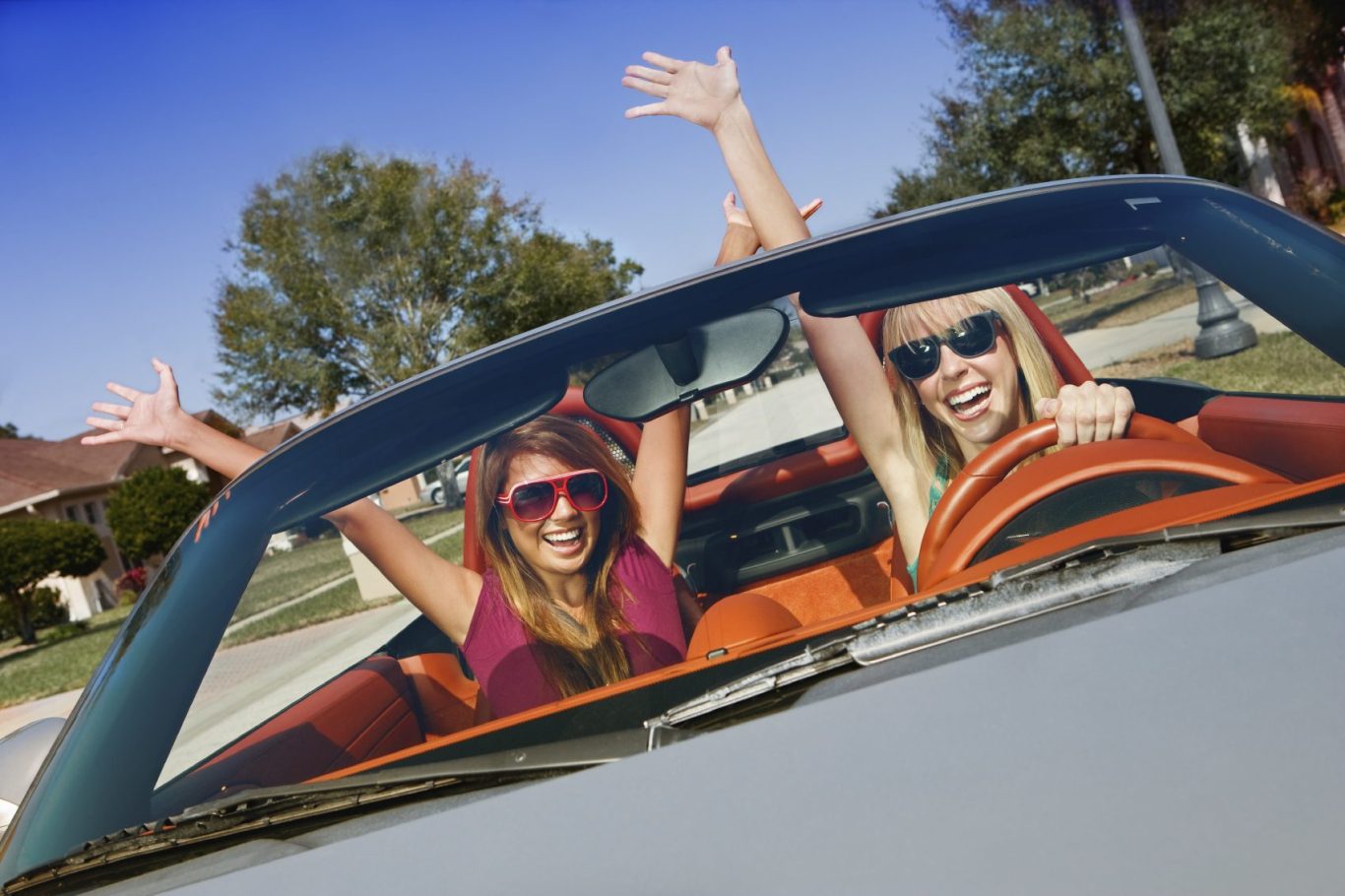 Zwei fröhliche Frauen in einem Cabrio, mit erhobenen Händen und Sonnenbrillen.