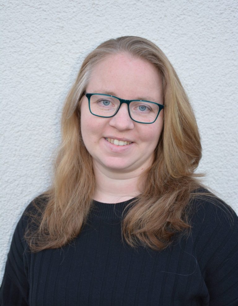 Lächelnde Frau mit langen, blonden Haaren und Brille, trägt einen schwarzen Pullover.
