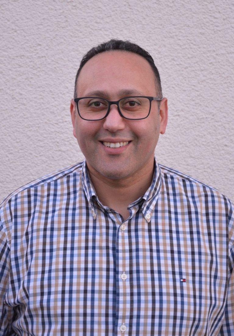 Lächelnder Mann mit Brille, trägt ein kariertes Hemd, vor einer hellen Wand.